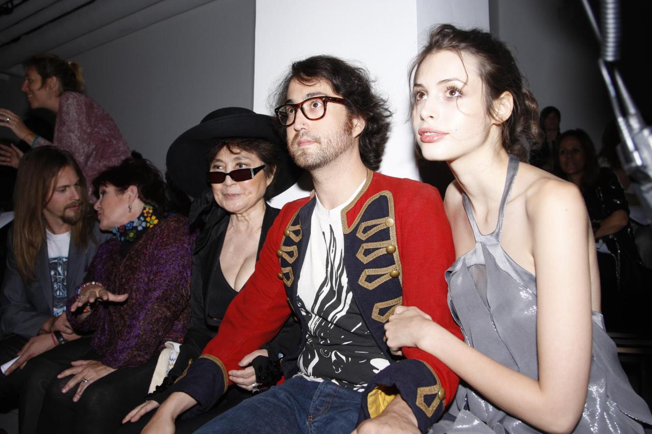 Sean Ono Lennon wears a red jacket with gold embroidered details while seated next to a woman in a gray dress at what appears to be an indoor event, with other guests visible in the background.