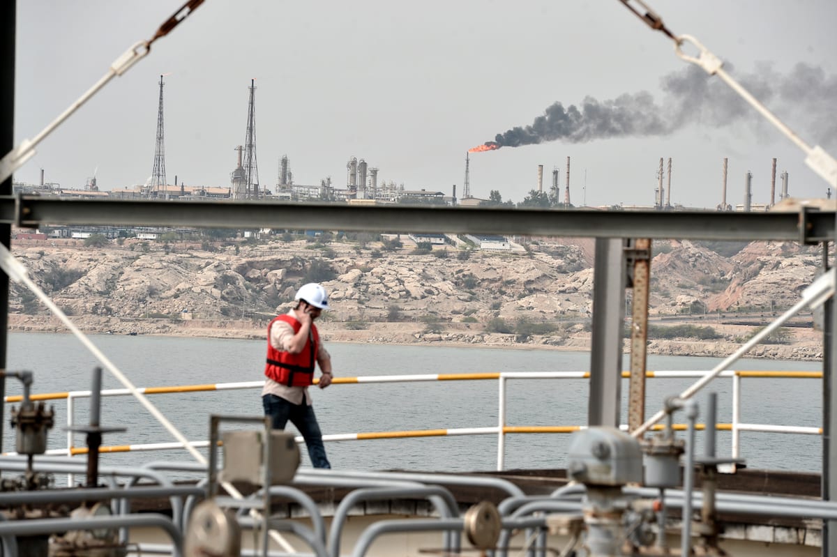 A worker in a red safety vest and hard hat stands on an industrial vessel deck overlooking a large oil refinery complex across the water, with smoke visible from industrial operations.