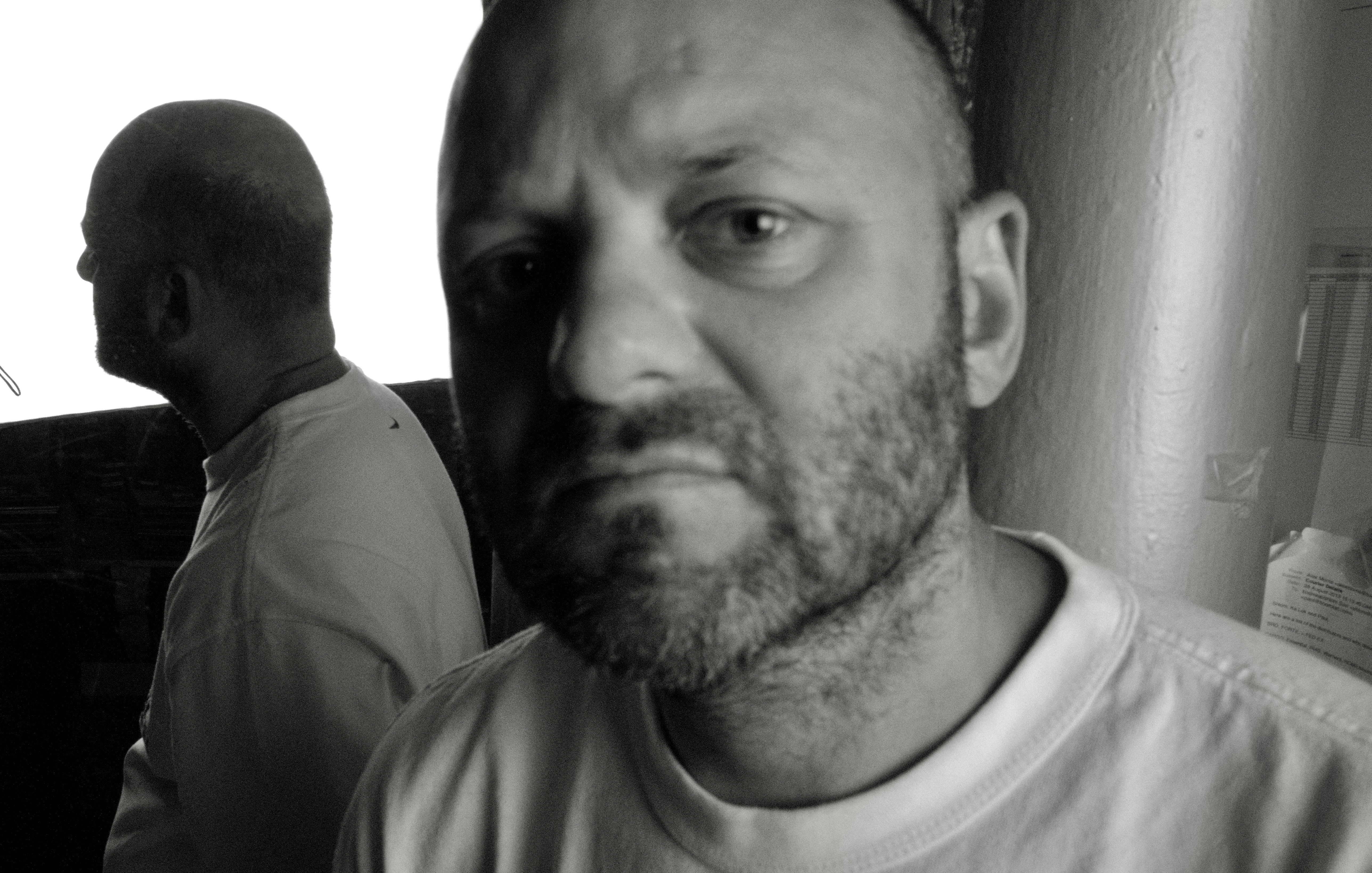 A black and white photograph of two men indoors; one man with a beard is in sharp focus in the foreground looking toward the camera, while another bald man is out of focus in the background.