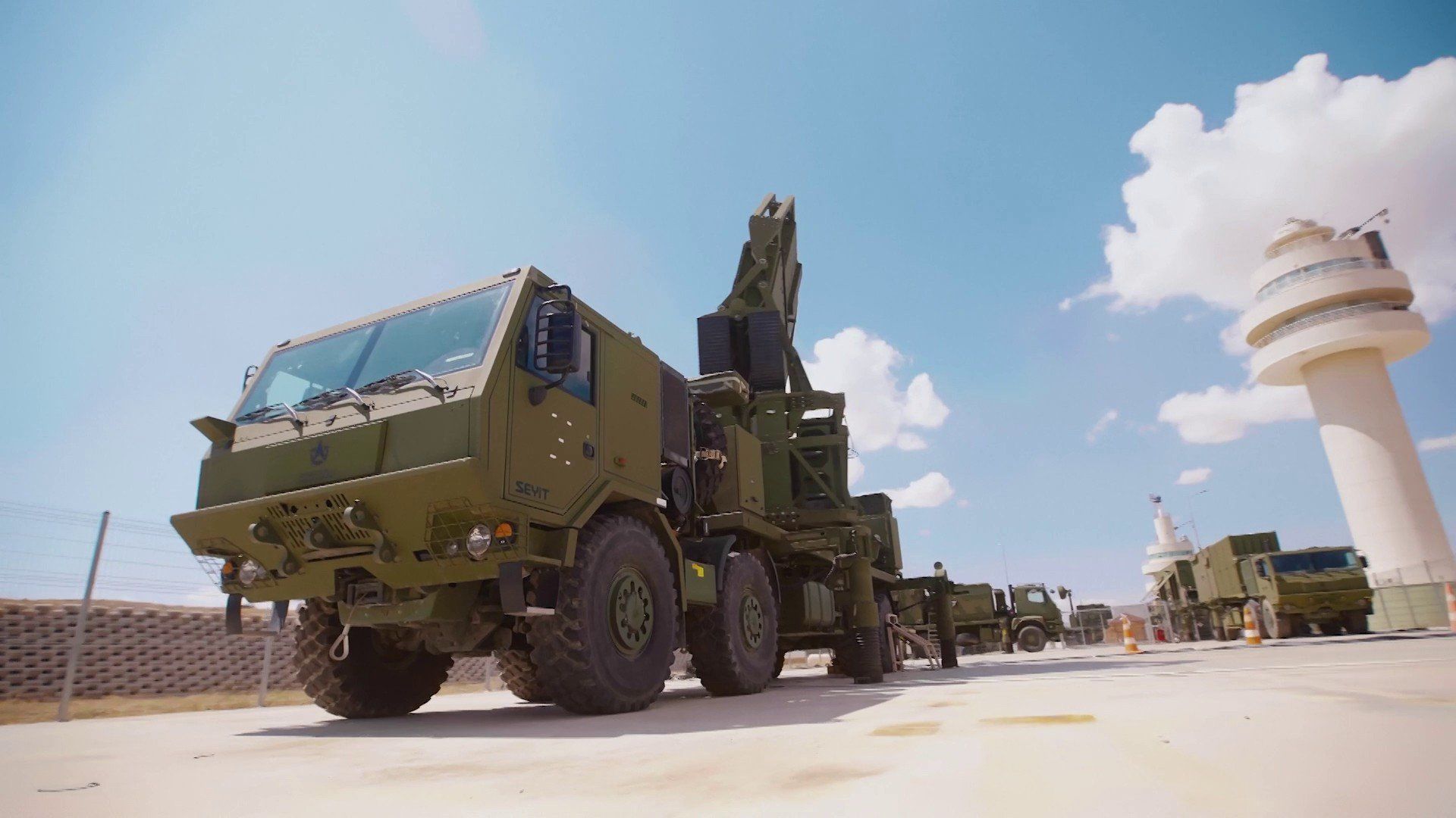 A military truck with olive-green coloring carrying radar and antenna equipment sits parked at what appears to be a military installation, with a control tower visible in the background.