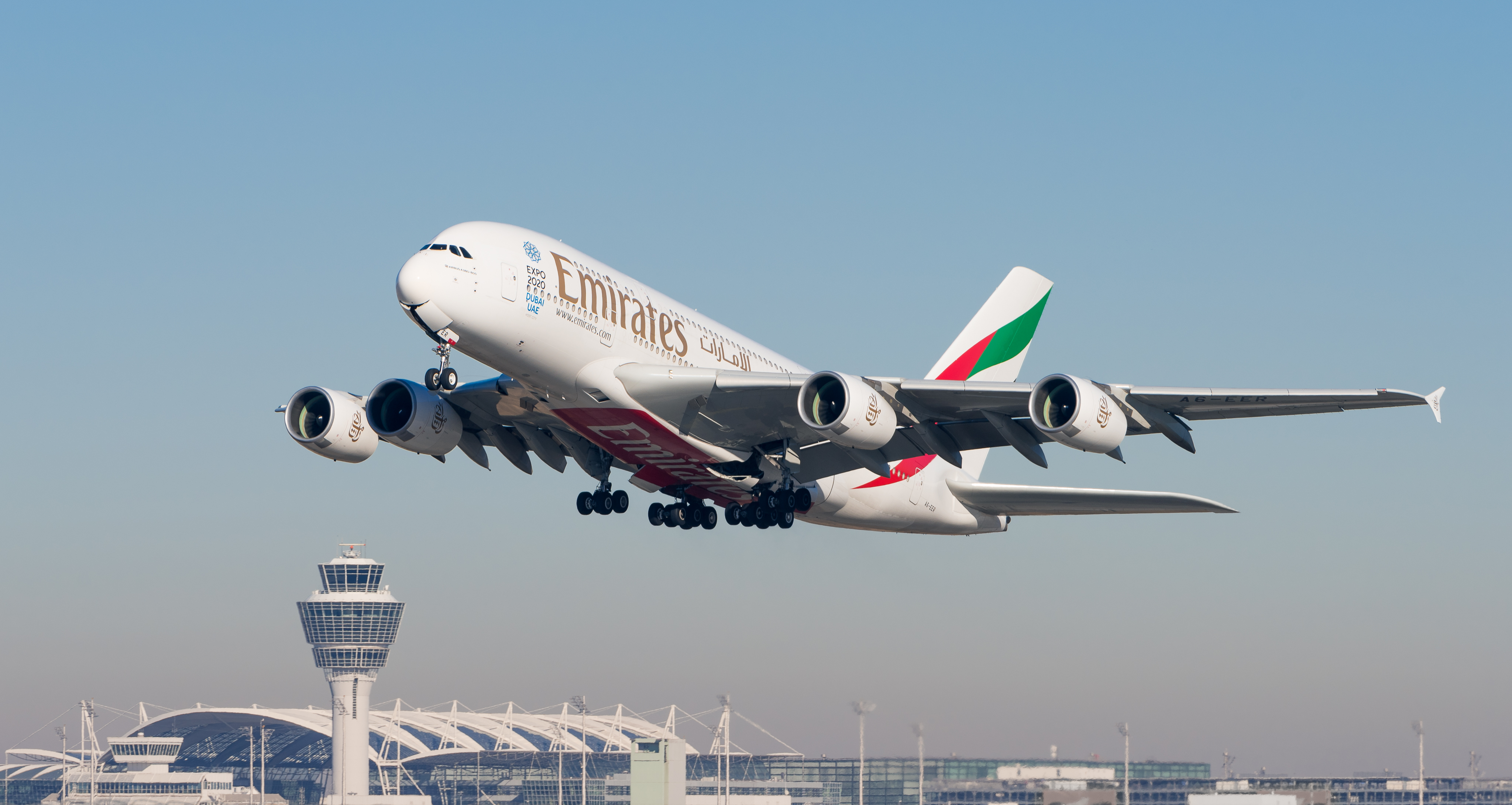 An Emirates airline Airbus A380 in flight taking off or approaching, with its landing gear deployed, shown against a clear blue sky with an airport control tower and terminal buildings visible below.