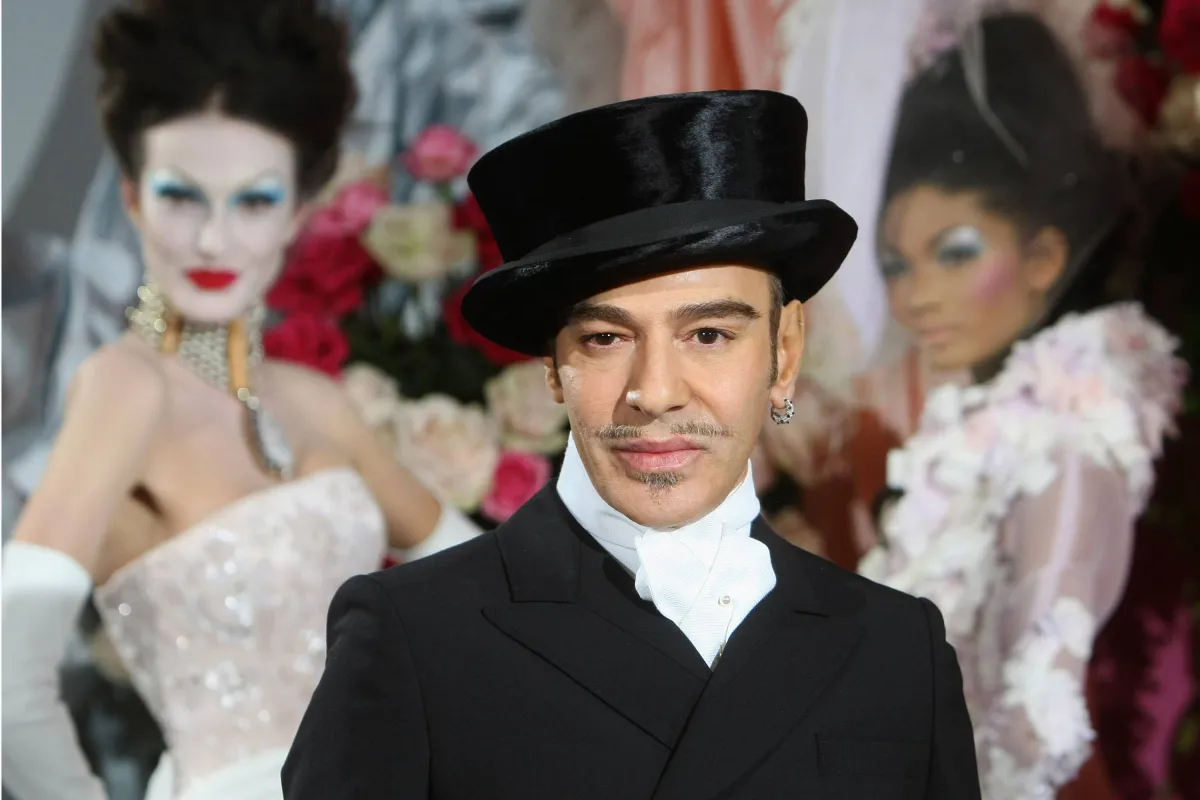 A man wearing a black velvet top hat and formal black tailcoat with white shirt stands in the foreground of what appears to be a high fashion show setting, with elaborately dressed models in the blurred background.