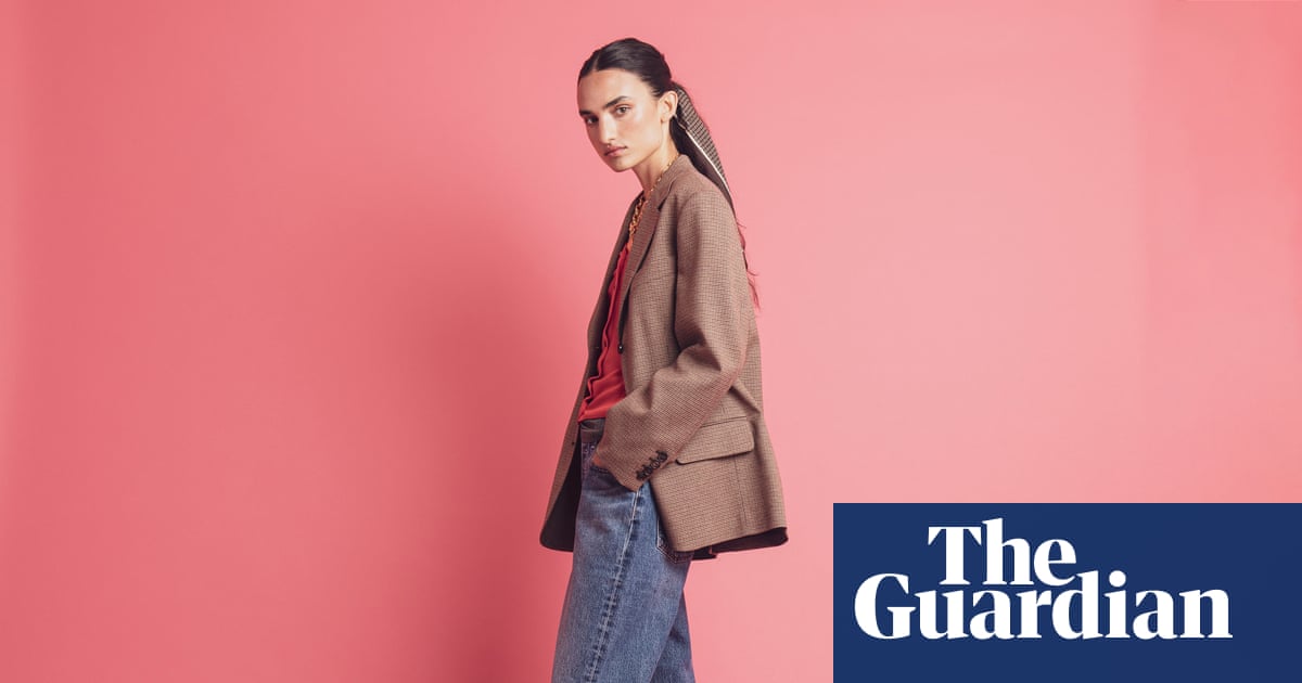 A woman poses against a pink background wearing a tan blazer over a red top and blue jeans.