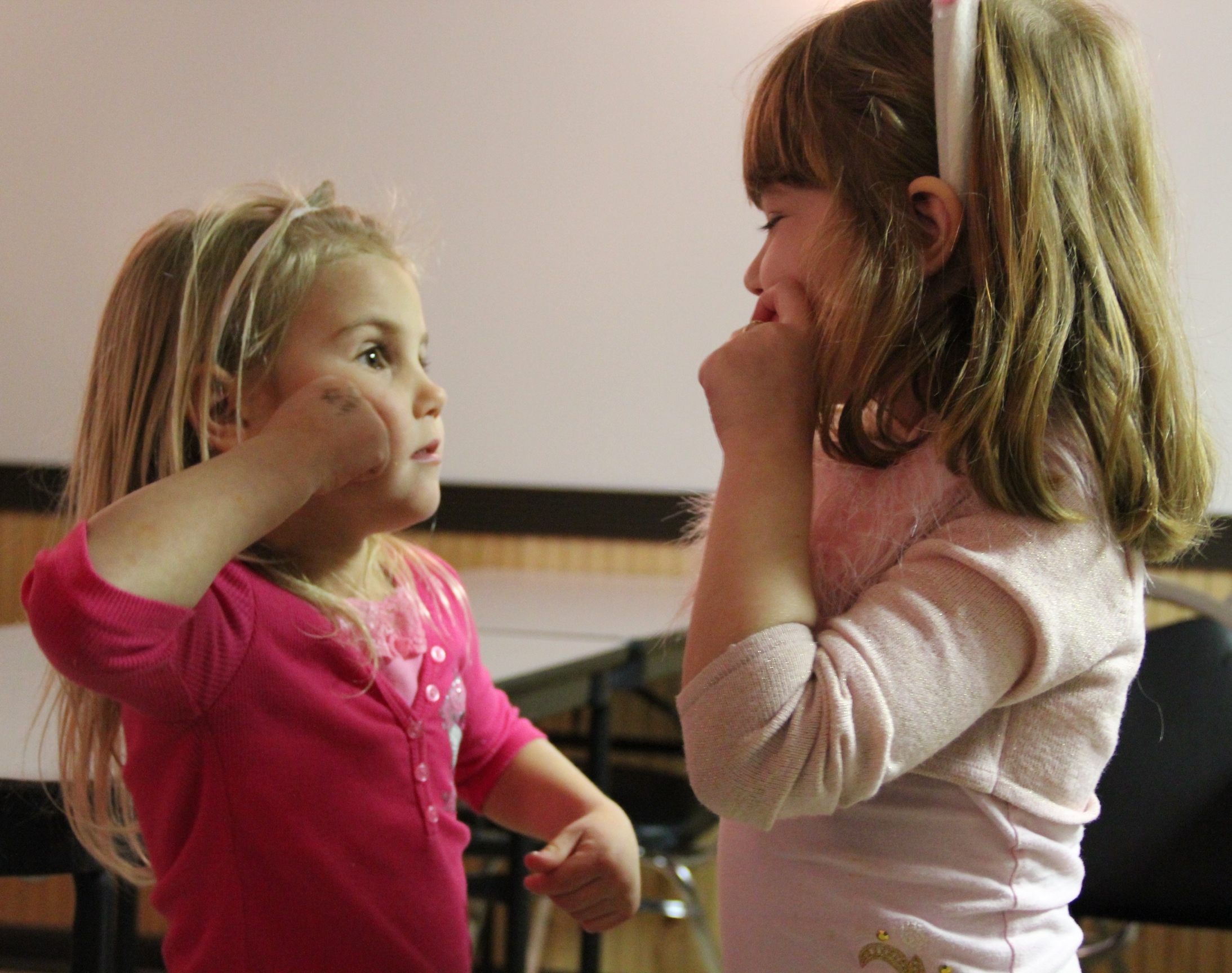 Two young girls face each other in an indoor setting, with one wearing a pink shirt pointing to her chin while looking at the other girl in a green shirt.