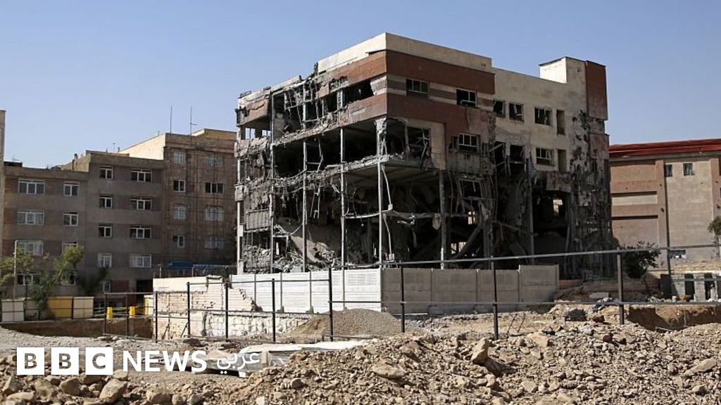 A heavily damaged multi-story brick building surrounded by rubble and a protective metal fence, with intact residential buildings visible nearby.