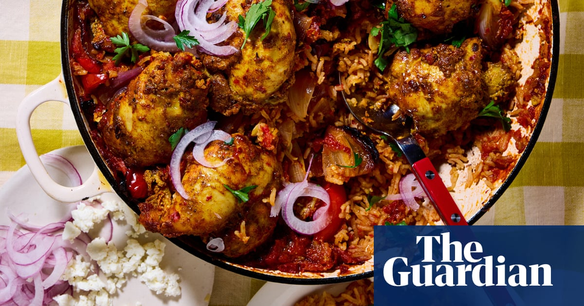 A large black paella pan filled with golden-brown spiced chicken pieces and rice, garnished with sliced red onions and fresh herbs.