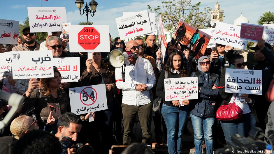 A large crowd of protesters holds signs in Arabic at a public demonstration, with historic architecture visible in the background.