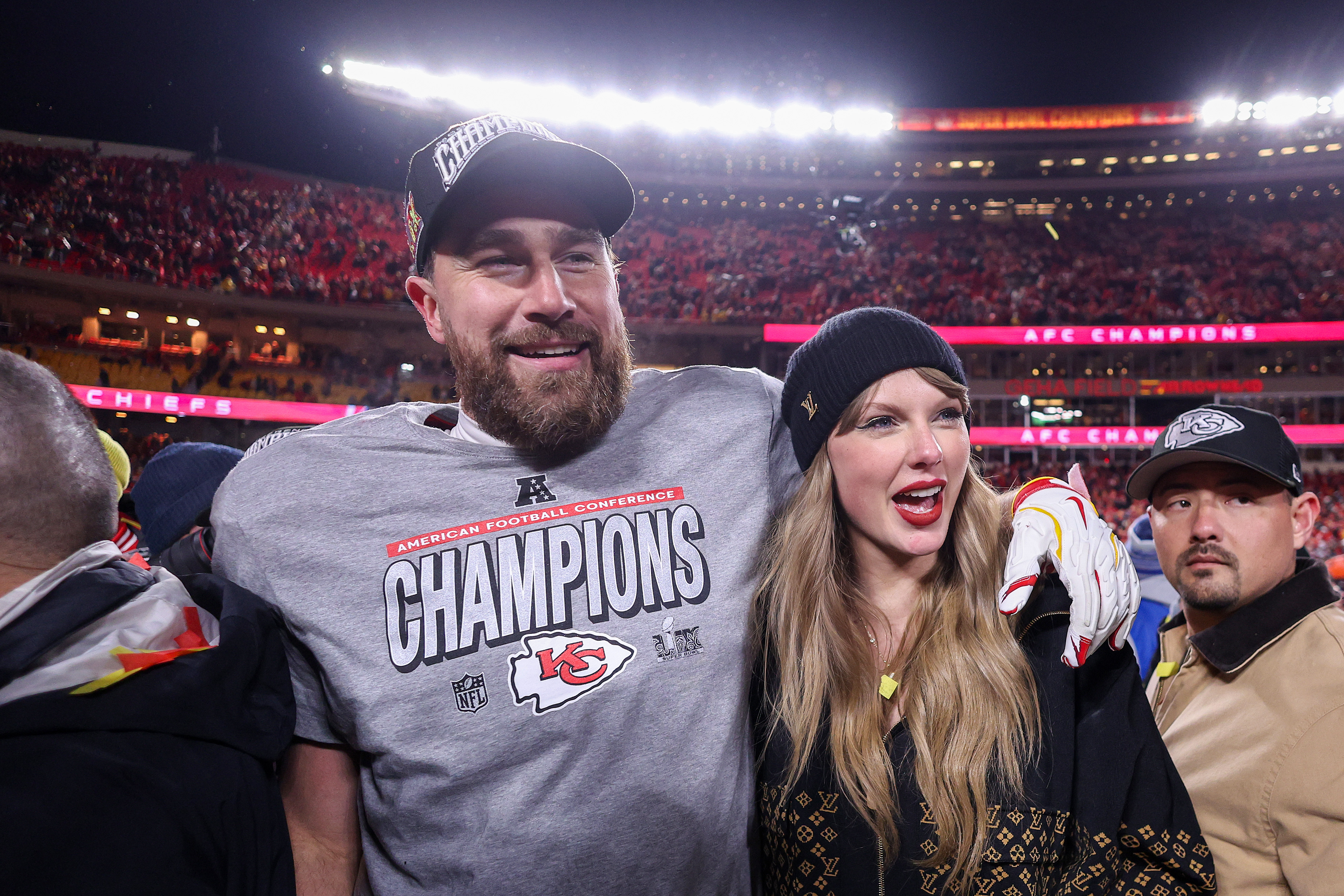 A man in a Kansas City Chiefs "AFC Champions" t-shirt and cap stands with a woman with long brown hair wearing a black beanie at a crowded NFL stadium.