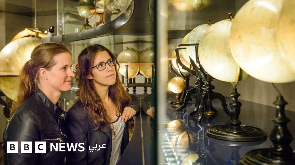 Two women stand in front of a glass display case examining antique globes illuminated by overhead lighting.