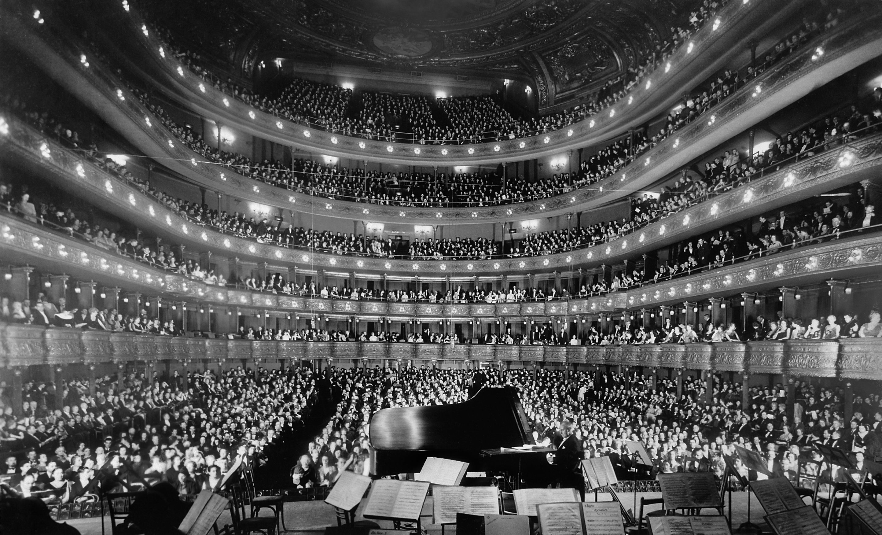 A grand concert hall filled with hundreds of audience members in balconies and seating areas, with a grand piano and music stands visible on the stage below.