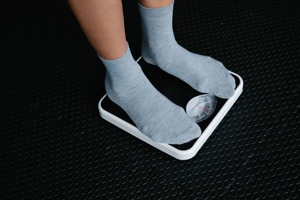 A person wearing gray socks stands on a white bathroom scale against a dark textured floor, with the scale's dial visible showing a weight measurement.