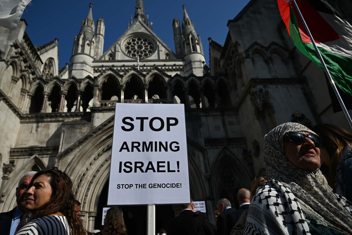 Protesters hold signs outside a Gothic cathedral, including one reading "STOP ARMING ISRAEL! STOP THE GENOCIDE" with Palestinian flags visible in the crowd.