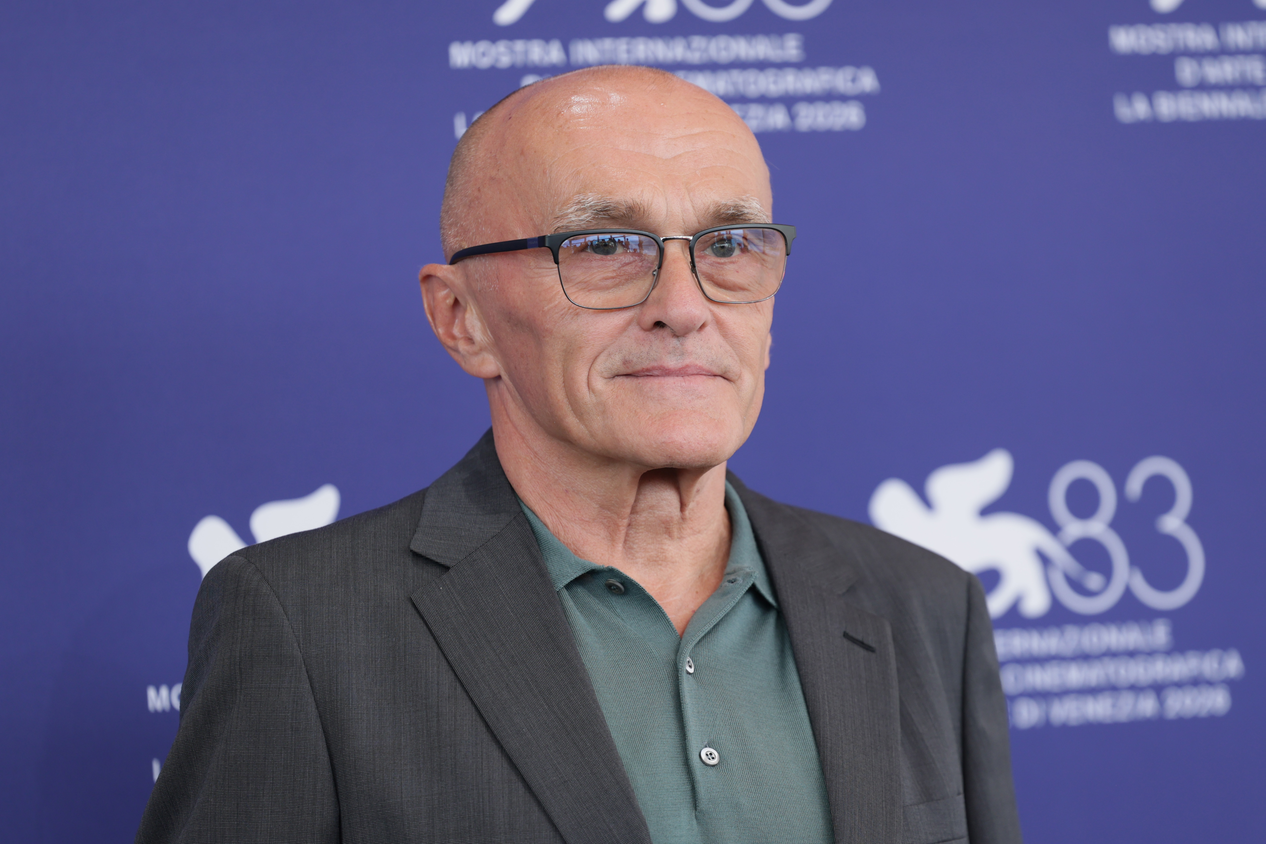 A man with glasses and a bald head wearing a gray blazer over a green shirt stands in front a blue backdrop with film festival signage.