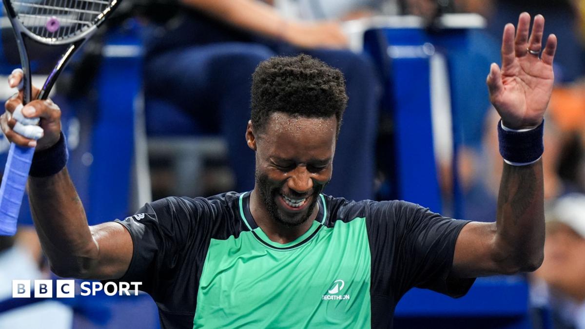 A tennis player celebrates with his arms raised and holding his racket, smiling broadly while wearing a green and black shirt and a wristband.