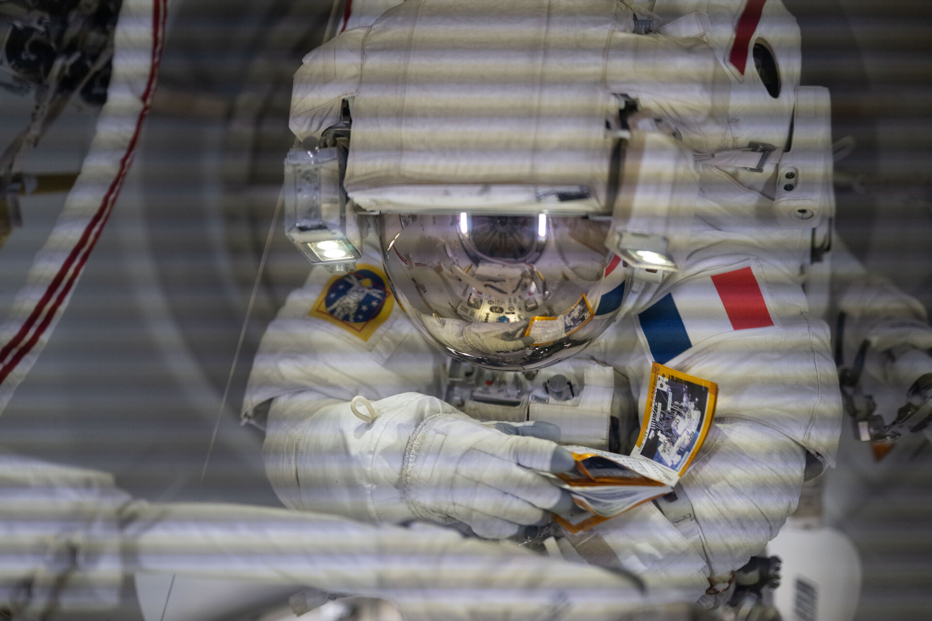 An astronaut in a white spacesuit performs a spacewalk outside the International Space Station, holding equipment and displaying a French flag patch on their suit.