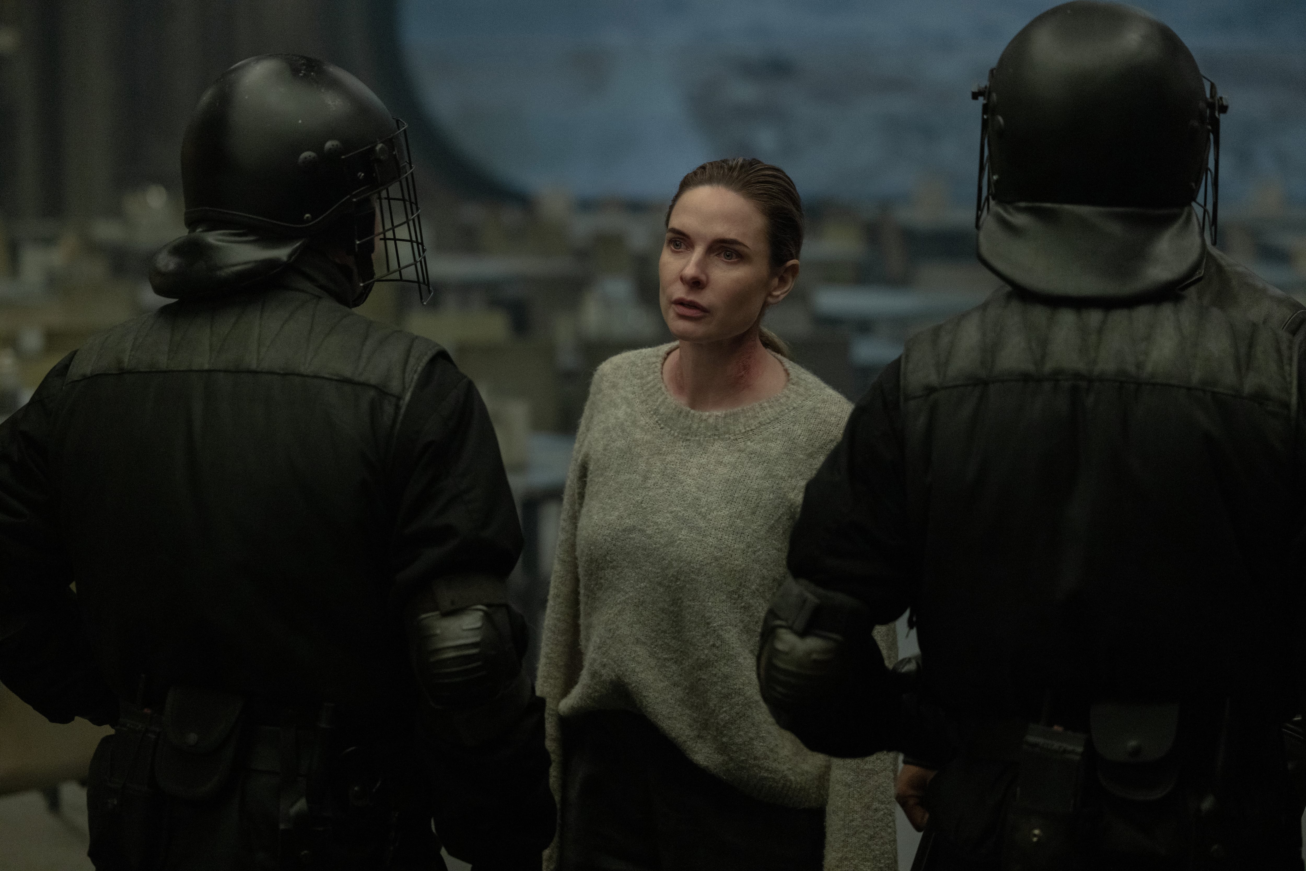 A woman in a beige sweater stands between two riot police officers wearing black helmets and tactical gear, with a blurred cityscape in the background.