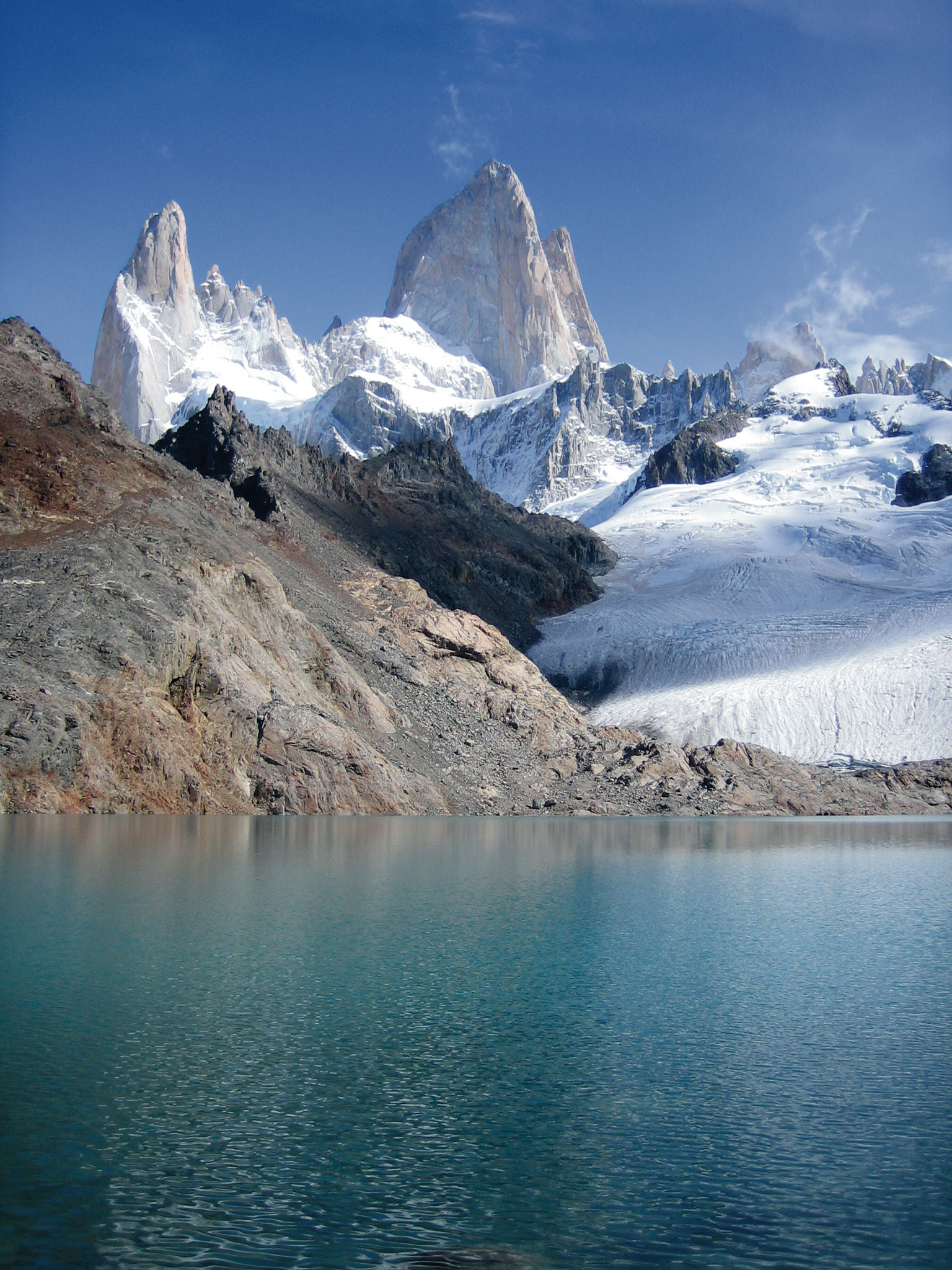 Snow-capped mountain peaks with jagged granite towers rise above a turquoise glacial lake, with bare rocky slopes in the foreground under a clear blue sky.