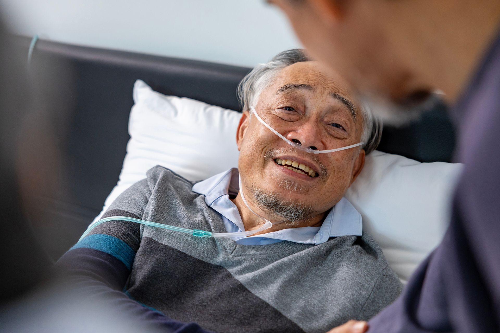 An elderly man with gray hair and a goatee smiles while lying in a hospital bed, wearing a light blue shirt and a nasal oxygen cannula, with another person partially visible in the foreground.