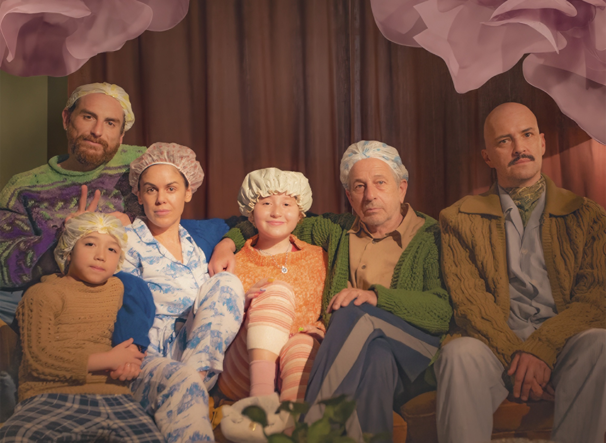 Six people of varying ages wearing colorful sweaters and sleeping caps sit together on a couch against a brown curtain backdrop with pink flower decorations.
