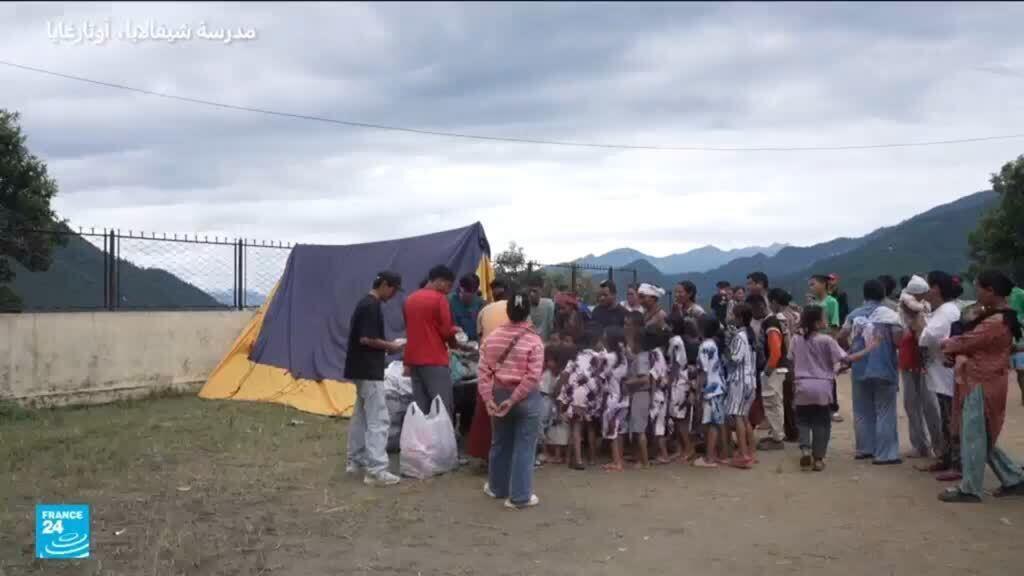 A large group of people, including many children, gather outside a blue and yellow tent in a temporary camp, with mountains visible in the background.